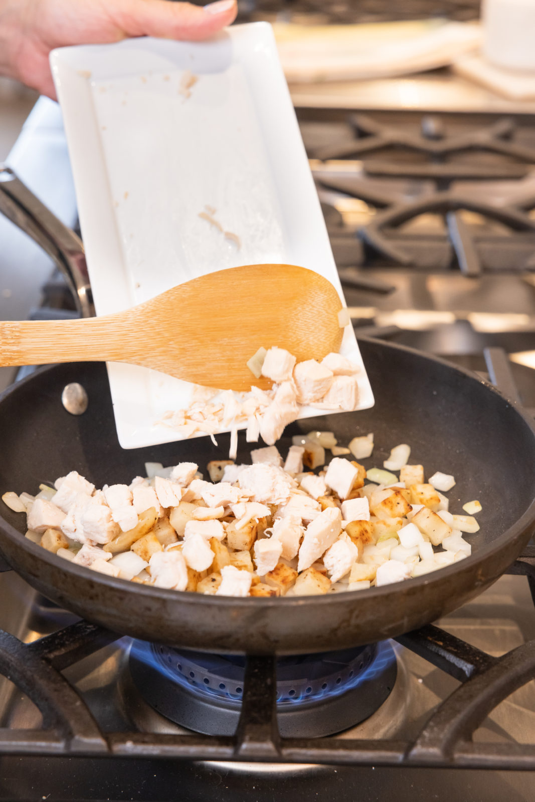 In this image, diced chicken and chopped onions are being added from a white dish to a sizzling pan on a gas stove, using a wooden spoon. The focus is on the action of combining these fresh ingredients, highlighting a step in the cooking process of a hearty meal.