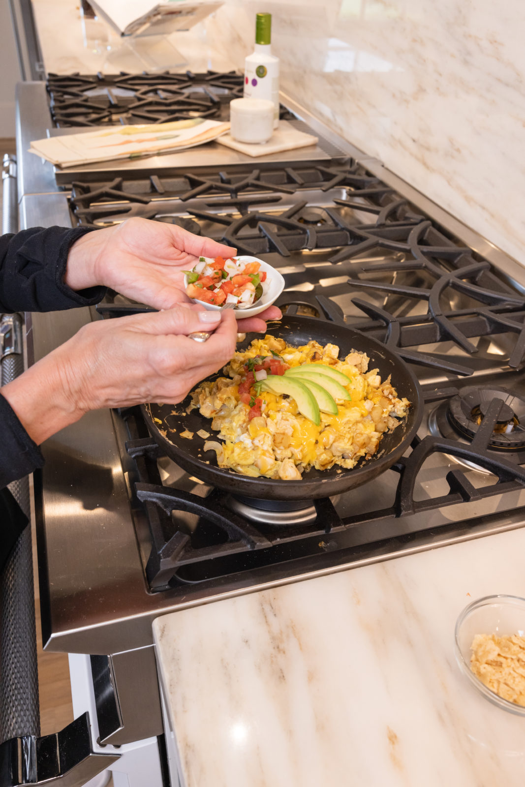 Hands are shown adding a small bowl of diced tomatoes and green peppers to a skillet with a colorful egg scramble topped with slices of avocado. The stove is set in a kitchen with a luxurious marble countertop and a sophisticated design.