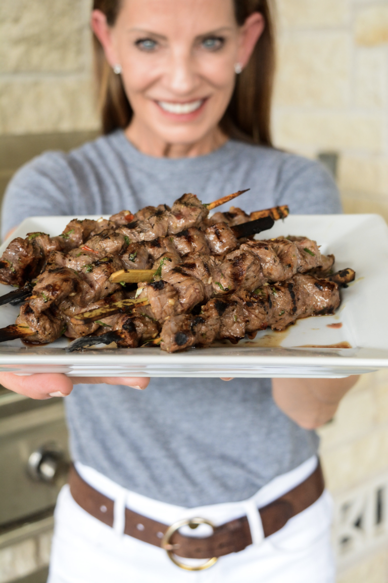 A woman smiling and holding a platter of grilled beef skewers garnished with herbs, presented in an outdoor setting.