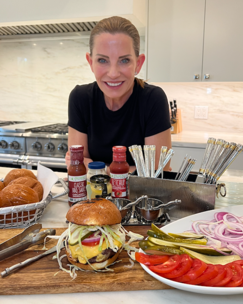 A woman with a joyful smile leans over a kitchen counter displaying a gourmet burger topped with cheese, lettuce, and tomato on a brioche bun, accompanied by condiments, fresh tomato slices, pickles, and sliced red onions.