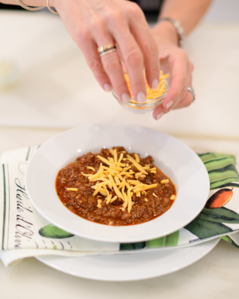 A person sprinkling shredded cheddar cheese over a bowl of hearty chili, served on a white plate with a decorative napkin on the side, capturing a moment of preparation in a bright kitchen setting.