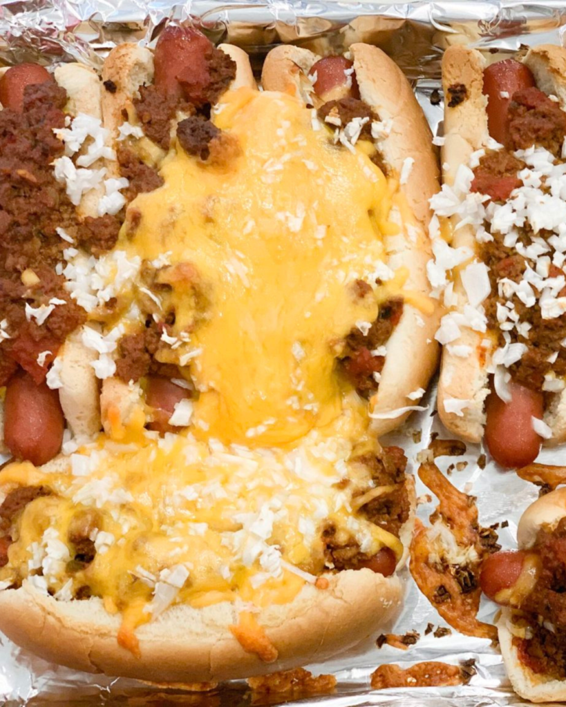 A tray of chili dogs topped with generous amounts of chili, shredded cheese, and onions, served in soft buns and cooked to golden perfection.