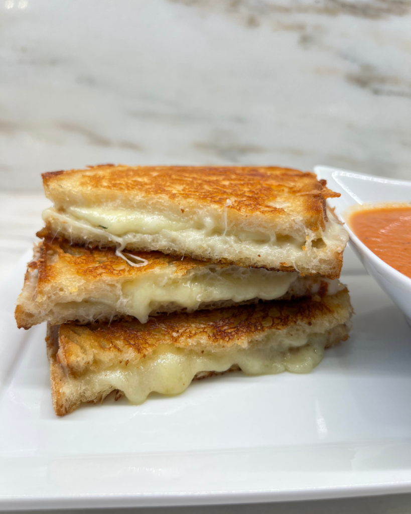 A close-up of a stack of three grilled cheese sandwich halves on a white plate. The sandwiches have golden-brown, crispy bread with melted cheese oozing out from the edges. The gooey cheese stretches slightly, emphasizing the rich texture. Part of a bowl of tomato soup is visible on the right side of the frame. The background features a blurred marble surface.