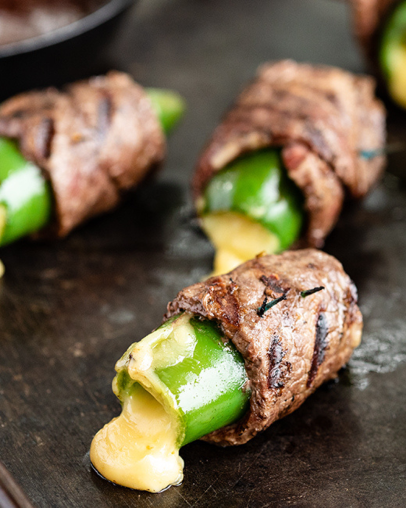 Grilled jalapeño poppers wrapped in seared steak, oozing melted cheese, placed on a dark cooking surface.