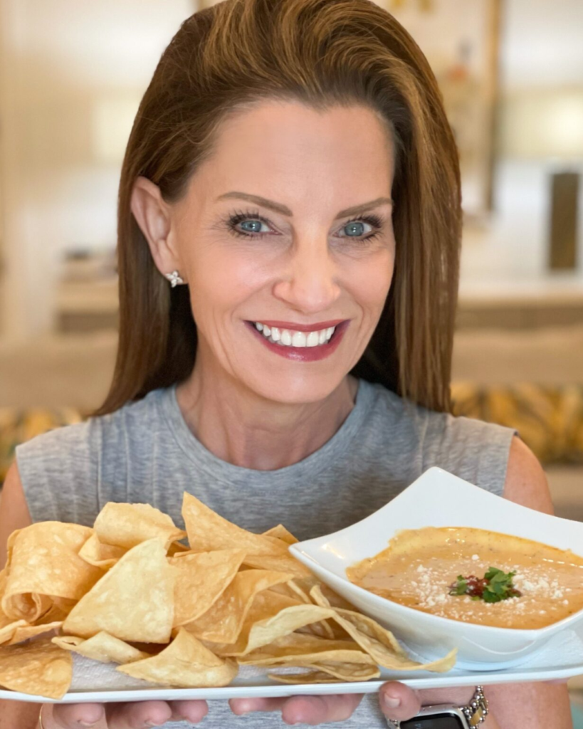 A woman with a radiant smile presenting a platter of crispy tortilla chips next to a bowl of creamy queso dip, garnished with fresh cilantro and crumbled cheese.