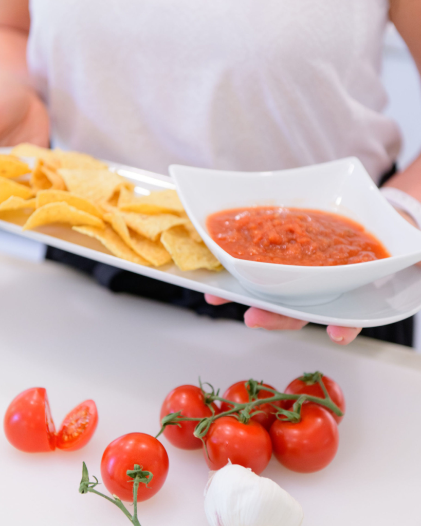 A close-up view of a person holding a serving tray with tortilla chips and a bowl of vibrant red salsa, surrounded by fresh ingredients like tomatoes, garlic, and cherry tomatoes on the vine.