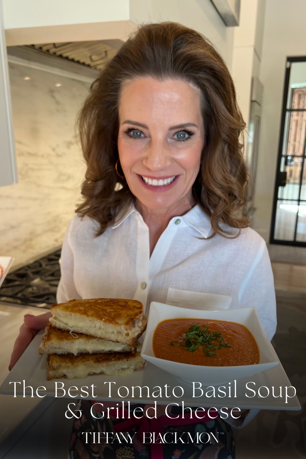 A smiling woman with shoulder-length wavy brown hair, wearing a white button-up shirt and a floral apron with a pink ribbon, holds a square white plate with a meal. The plate contains a grilled cheese sandwich, cut into halves with melted cheese oozing out, stacked neatly on one side, and a bowl of tomato soup garnished with fresh green herbs on the other. She stands in a bright, modern kitchen with a marble backsplash and a gas stove in the background