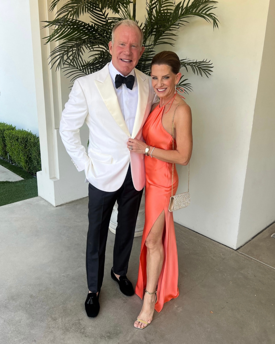 A man and a woman posing together at an outdoor event. The man is wearing a white dinner jacket with a black bow tie and black trousers, and the woman is in an elegant coral gown with a high slit, complemented by a beige clutch and gold strappy sandals. They are standing in front of a white wall with potted palm plants, both smiling at the camera.