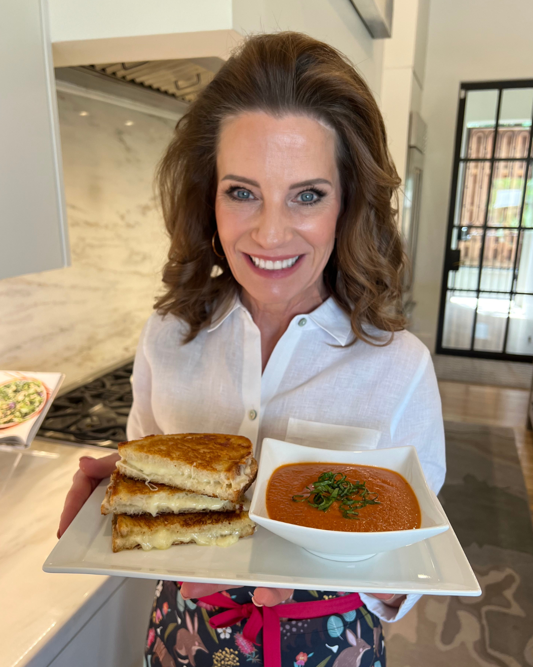 A smiling woman with shoulder-length wavy brown hair, wearing a white button-up shirt and a floral apron with a pink ribbon, holds a square white plate with a meal. The plate contains a grilled cheese sandwich, cut into halves with melted cheese oozing out, stacked neatly on one side, and a bowl of tomato soup garnished with fresh green herbs on the other. She stands in a bright, modern kitchen with a marble backsplash and a gas stove in the background