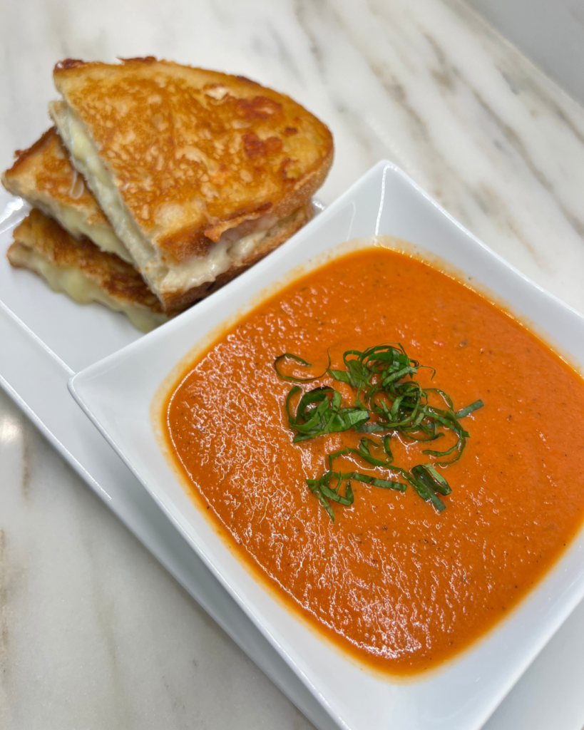 A close-up of a bowl of creamy tomato soup with a vibrant orange-red color, garnished with thinly sliced fresh basil. The soup is served in a square white bowl, which rests on a matching white plate. Beside the bowl, a stack of golden-brown grilled cheese sandwiches, cut into halves, is placed on the plate. The melted cheese is visible between the crispy slices of bread. The background features a marble countertop with a subtle pattern.