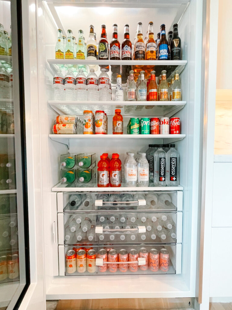 "A neatly organized refrigerator stocked with various bottled and canned beverages, including water, sparkling water, sports drinks, and soda. Clear storage bins keep drinks arranged, creating a tidy and aesthetically pleasing display.