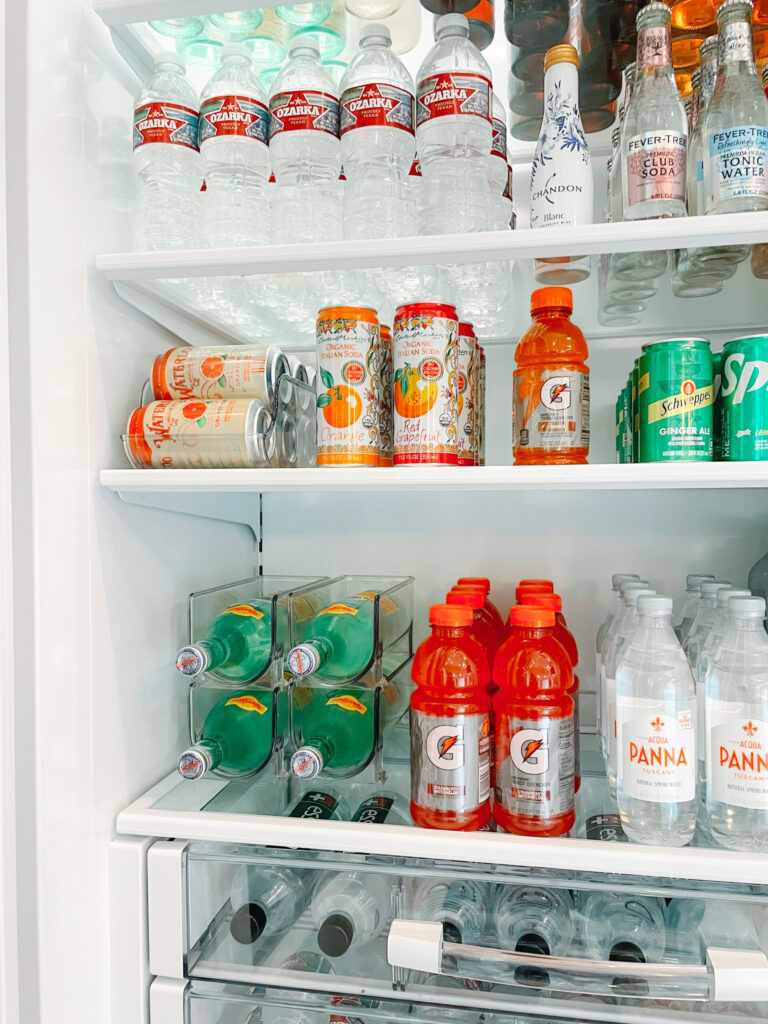 "A neatly organized refrigerator stocked with various bottled and canned beverages, including water, sparkling water, sports drinks, and soda. Clear storage bins keep drinks arranged, creating a tidy and aesthetically pleasing display.