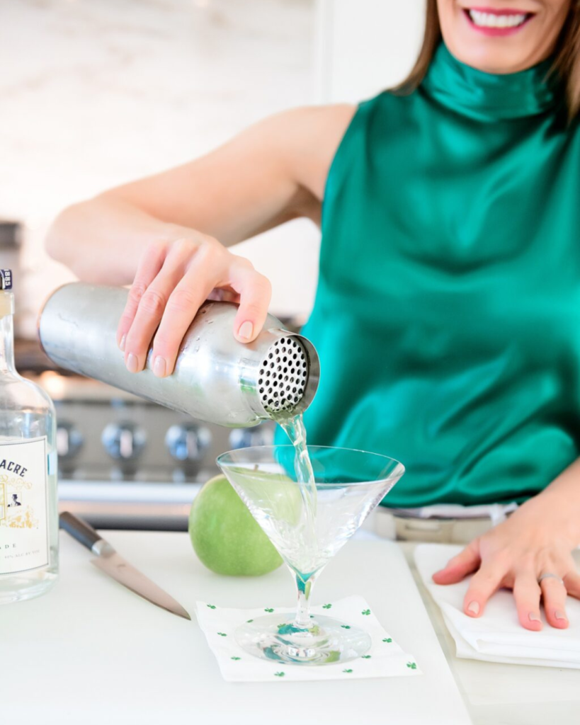 In this image, a woman in an elegant emerald green top is seen pouring a clear liquid from a cocktail shaker into a martini glass. Her cheerful expression and the casual elegance of her attire add a vibrant and welcoming atmosphere to the scene. The countertop holds a green apple and a knife, suggesting fresh ingredients are used in her cocktail preparation. A bottle labeled "Sour Acre" and a decorative napkin with a clover pattern are also visible, enhancing the theme of freshness and flavor.