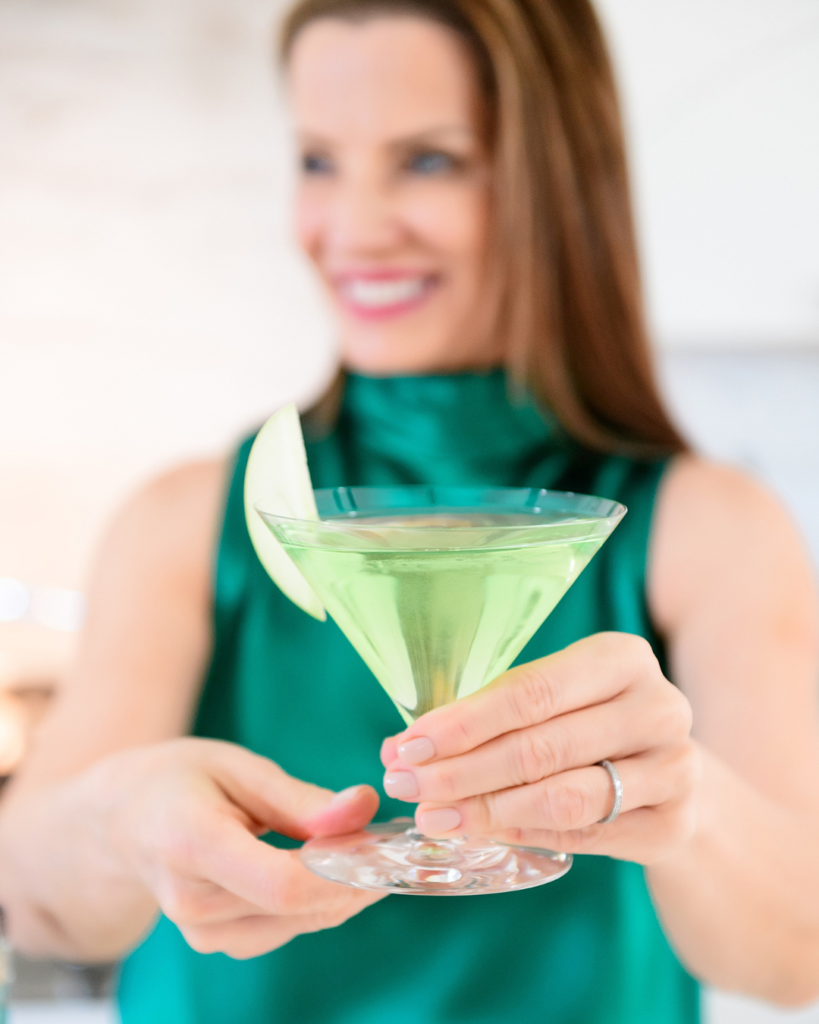 In this image, a woman with a radiant smile is offering a vibrant green cocktail in a martini glass. She is wearing an elegant emerald green top that complements the color of the drink. The focus is on the woman and the cocktail, with a softly blurred background enhancing the intimate and cheerful ambiance. A slice of apple garnishes the drink, adding a fresh touch to the scene.